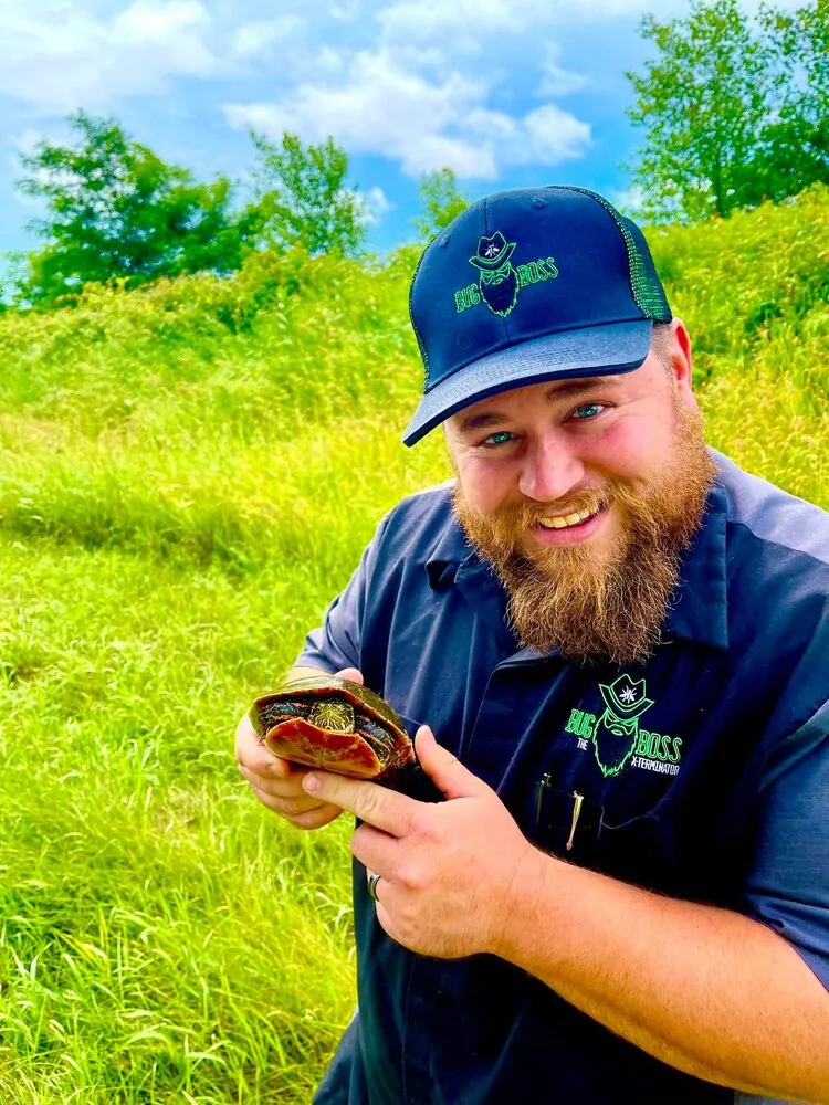 Brian Zahringer, owner of BugBoss The X-Terminator, in a branded shirt and cap holding a painted turtle in a rural Wisconsin field